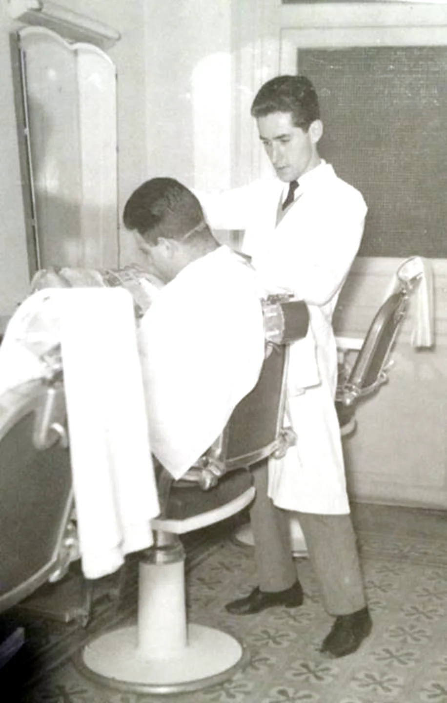 A young barber in a white coat and tie cutting a client's hair at Madison's